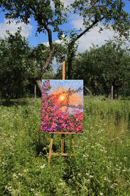 A wooden easel stands in a sunlit field of green grass and white daisies, holding a vibrant painting of a colorful sunset…