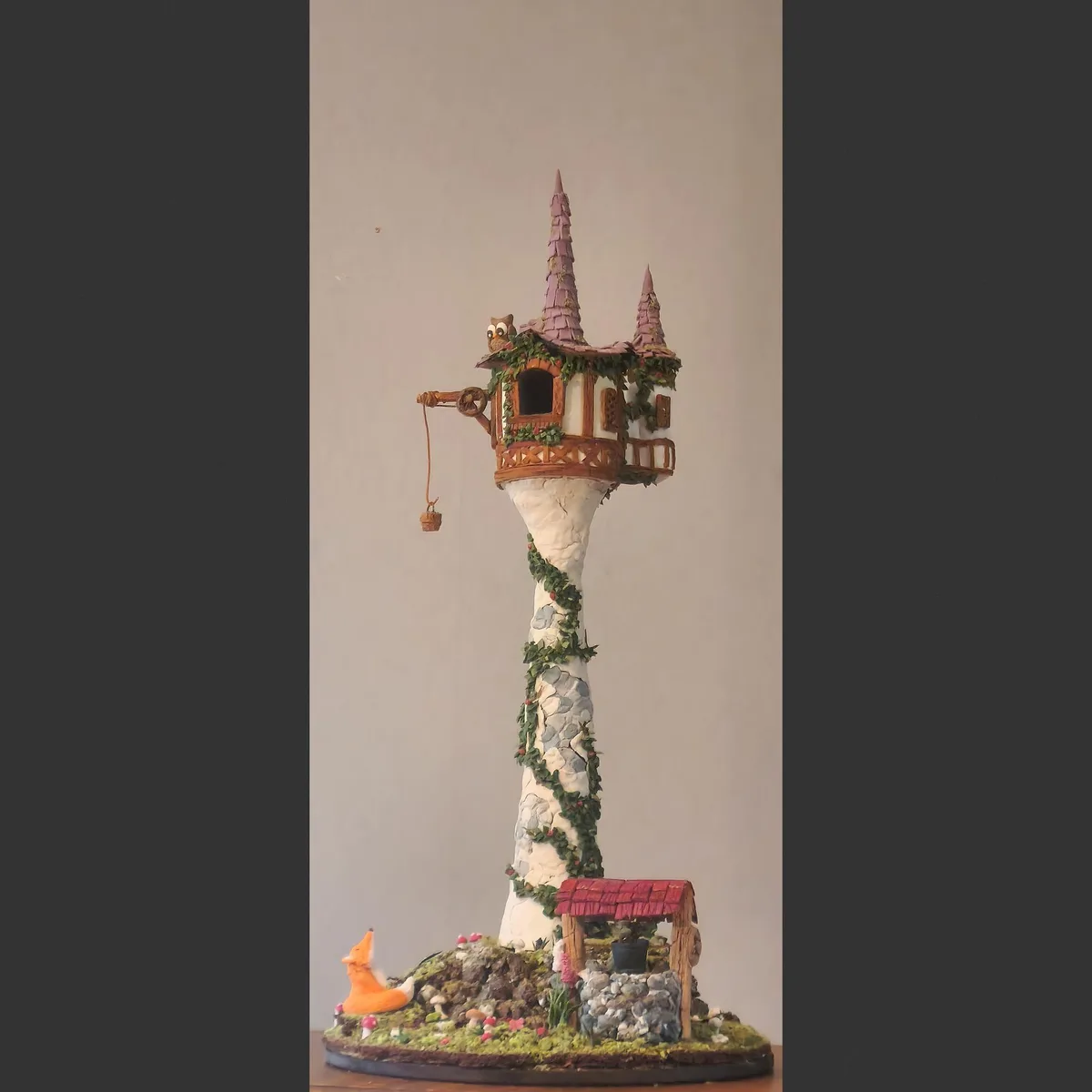 A handmade clay model of Rapunzel's tower from Disney's Tangled, featuring a tall stone structure with ivy, a balcony, a…