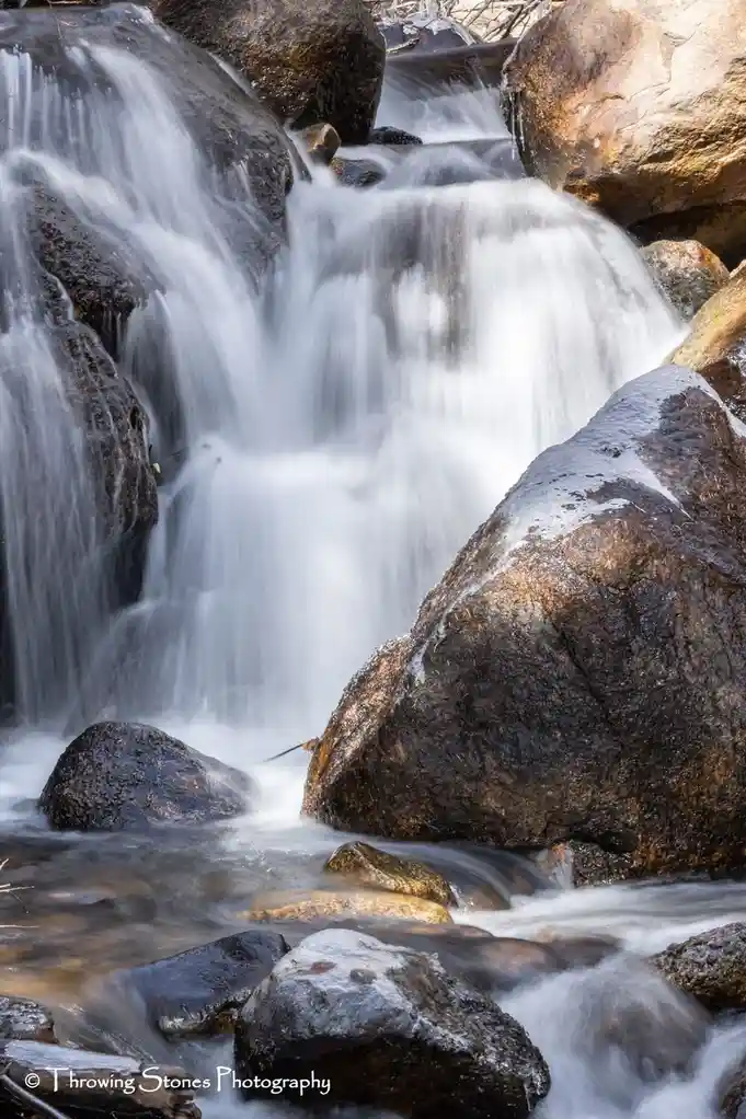 A long-exposure photograph shows a serene, blurred waterfall cascading over dark, wet rocks with some ice patches. The…