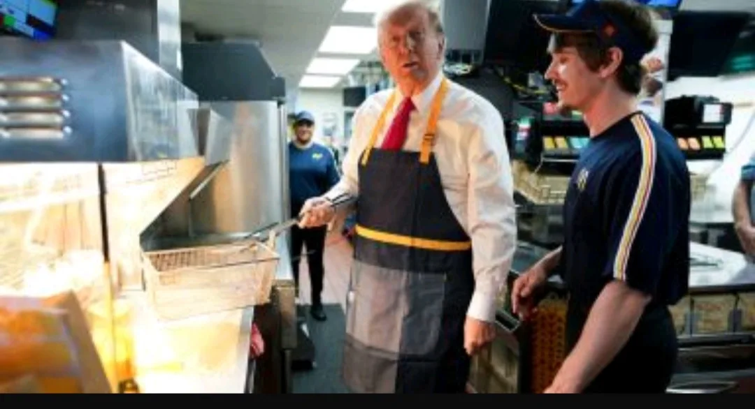 A man in a white shirt, red tie, and black apron with yellow straps stands in a fast-food kitchen holding a fry basket over…