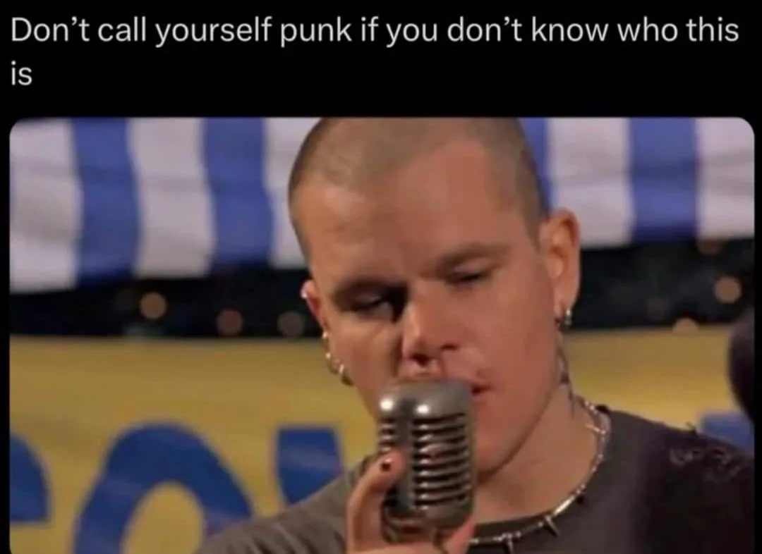 A close-up shot of a man with a shaved head, wearing multiple earrings and a spiked necklace, singing into a vintage-style…