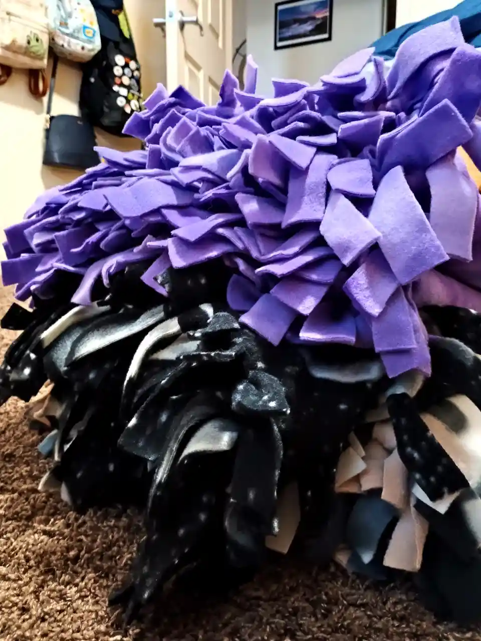 A close-up photo shows a handmade, multi-colored fleece snuffle mat, primarily purple and black, sitting on a brown carpet…