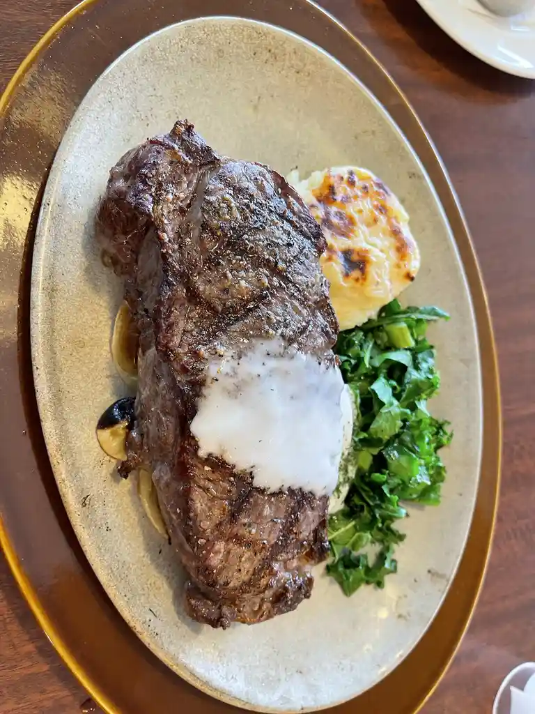A close-up photo of a plated steak with a large, white sauce blob covering its center, accompanied by roasted potatoes and…