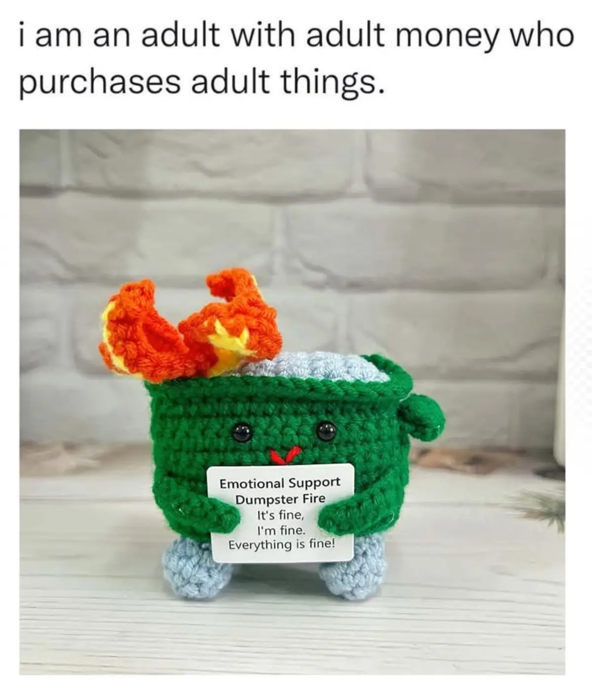 A green crocheted plushie resembling a dumpster fire with orange flames on top, holding a sign that reads 'Emotional Support…