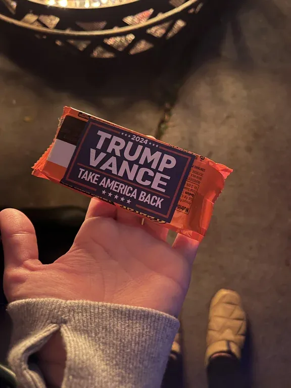 A hand holds a candy bar wrapper designed to look like a political campaign item, featuring the text '2024 TRUMP VANCE TAKE…