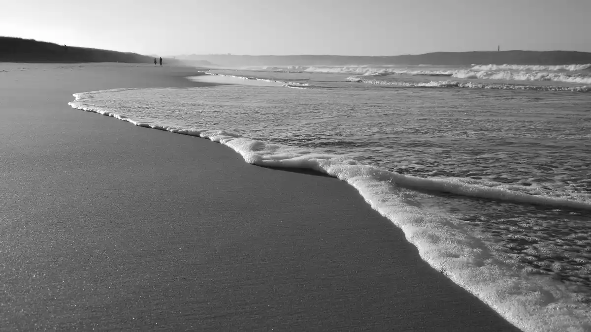 A black-and-white photo of a calm beach with gentle waves rolling onto the shore, two distant figures walking along the…