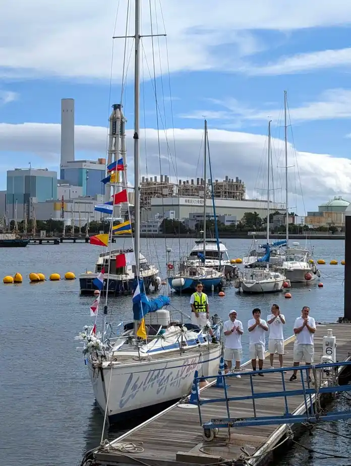 A group of people in white shirts stand on a dock clapping, with a sailboat in the foreground and a large nuclear power…