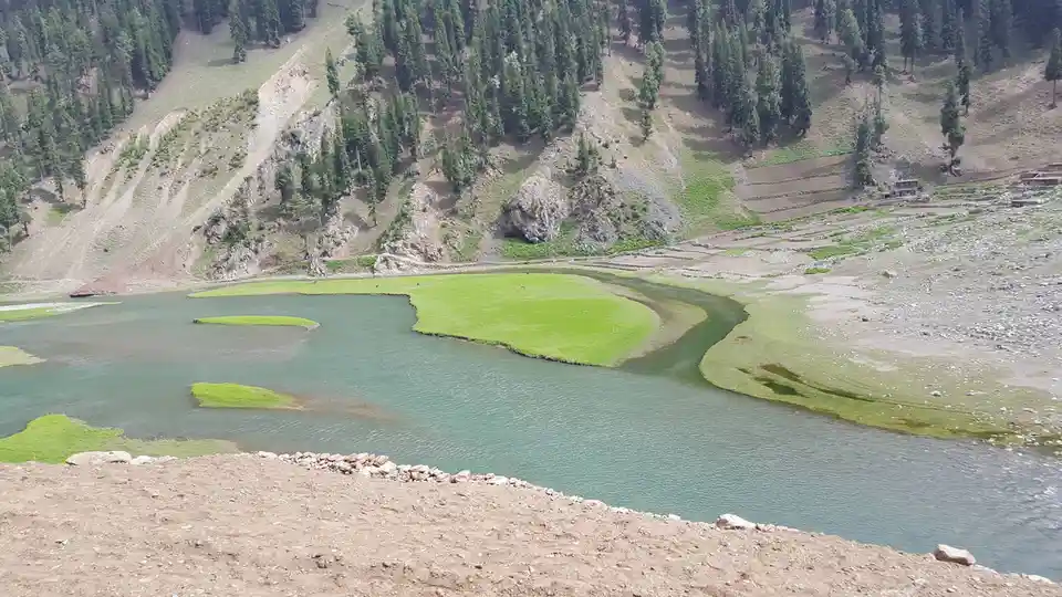 a scenic mountain valley with a river flowing around a vibrant green, heart-shaped grassy island in the middle of a…