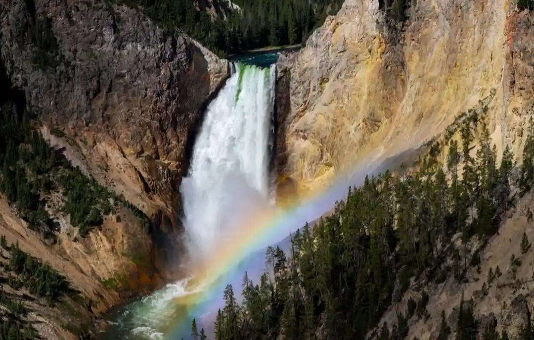 A majestic waterfall cascades down a rocky canyon, surrounded by dense evergreen forests, with a vibrant rainbow arcing…