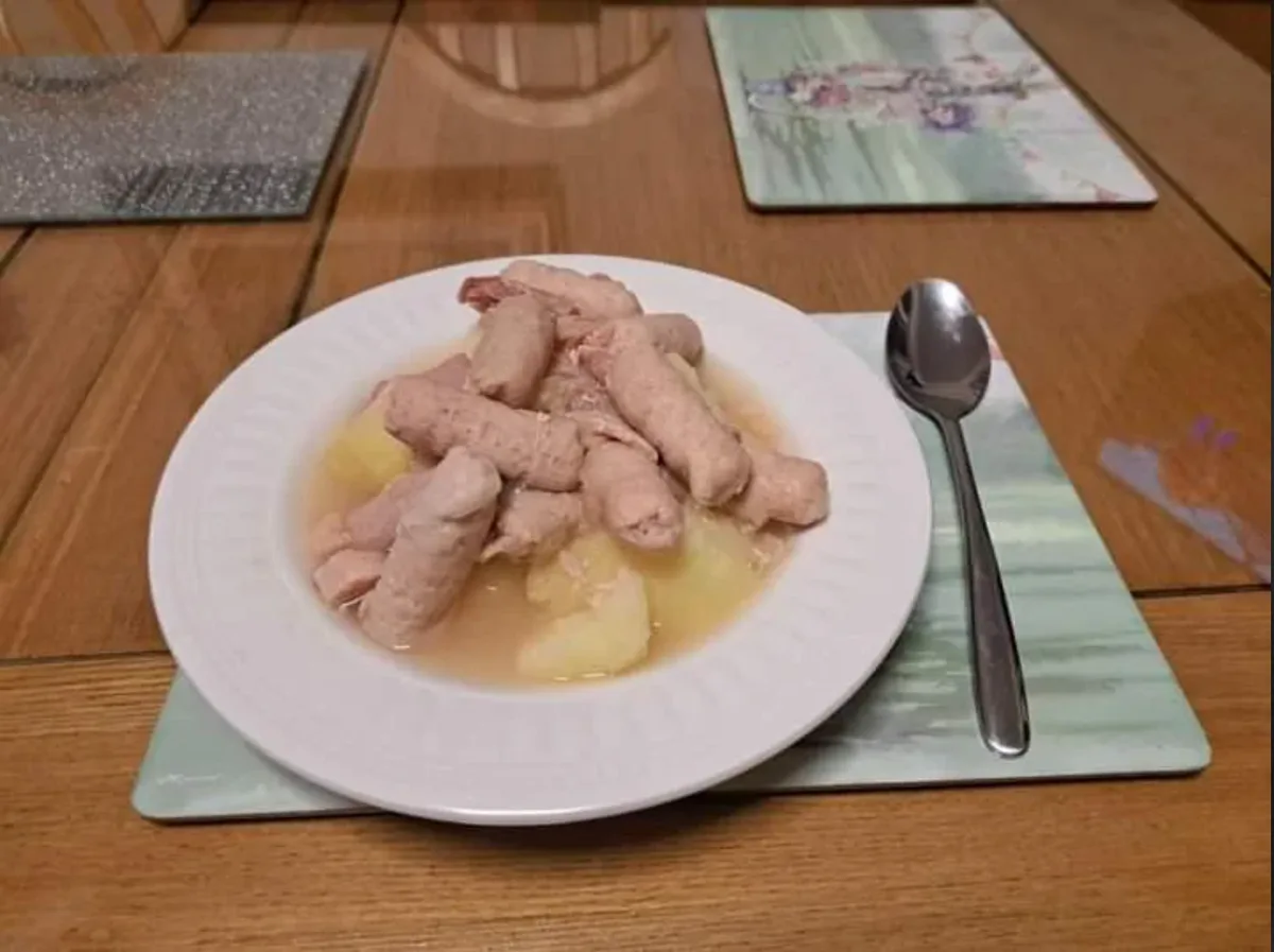 A white plate on a wooden table holds a serving of cooked sausages and potato chunks in a light broth, with a metal spoon…