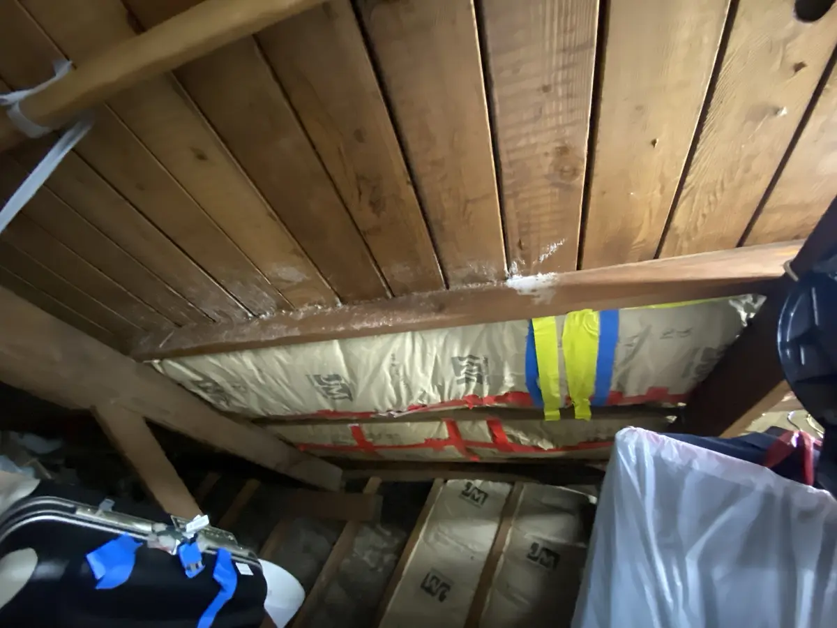 A tilted photo of a cluttered attic or crawl space with exposed wooden beams and insulation rolls. White insulation dust is…