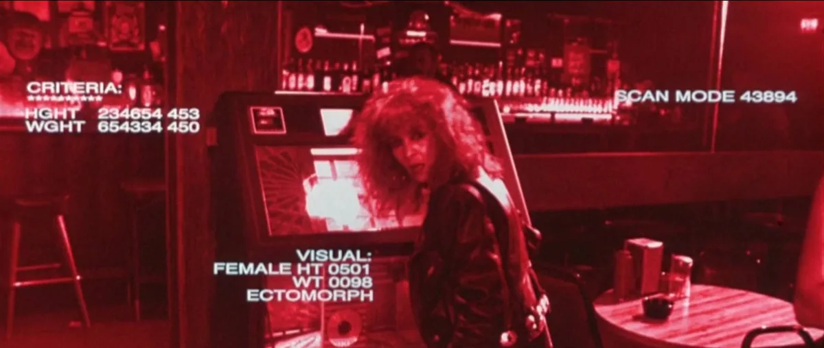 A woman with curly red hair and a leather jacket stands in a dimly lit bar, viewed through a red-tinted security camera…