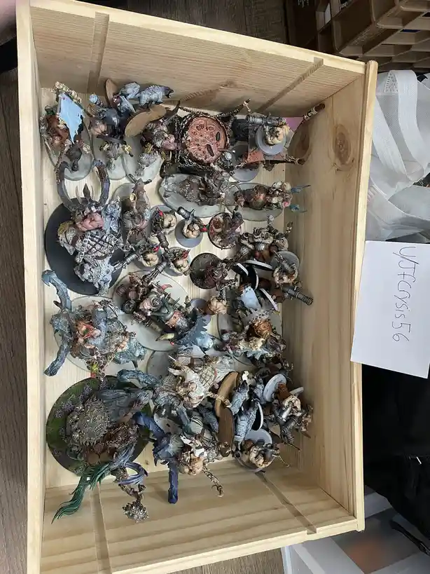 A wooden crate is filled with numerous painted miniature figurines, mostly depicting fantasy or warhammer-style warriors and…