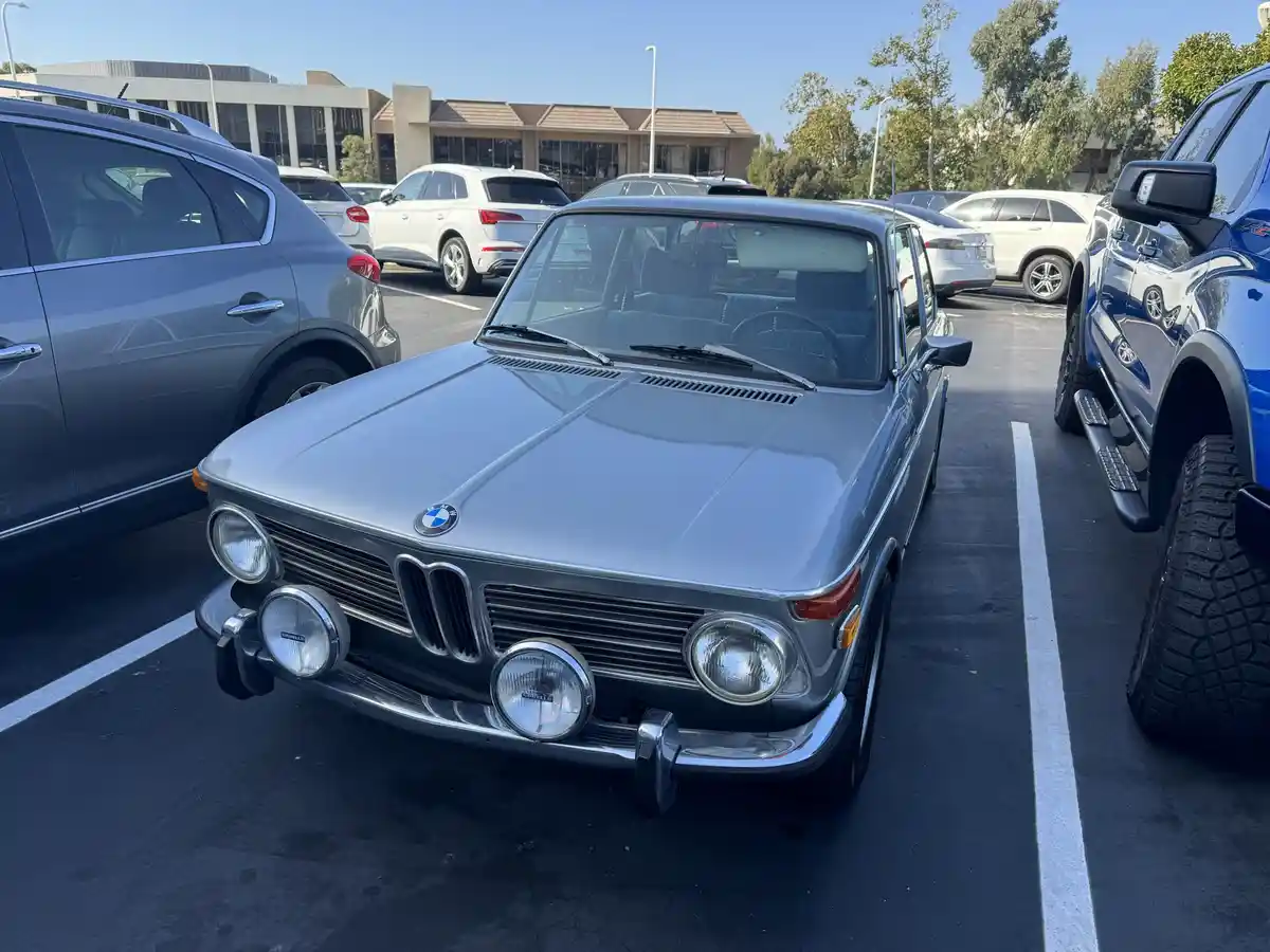 A silver vintage BMW sedan with round headlights and a chrome grille is parked in a modern parking lot surrounded by…
