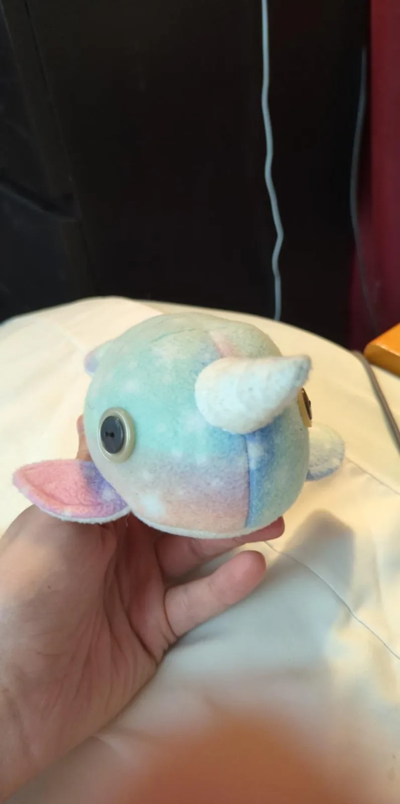 A hand holds a small, handmade plush toy resembling a narwhal, featuring pastel blue, pink, and white fabric, a single white…
