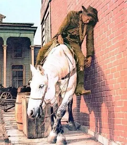 A man in old western attire, wearing a hat and boots, is slumped against a red brick wall while sitting atop a white horse…