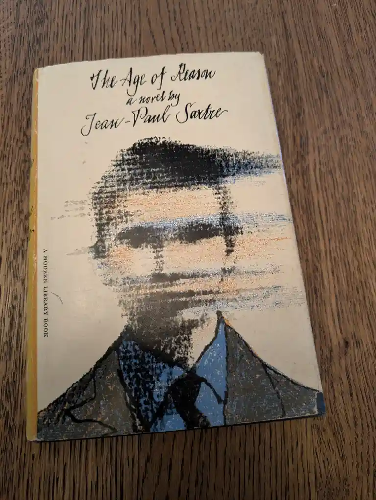 A close-up photo of a vintage paperback book titled 'The Age of Reason' by Jean-Paul Sartre, resting on a wooden surface…