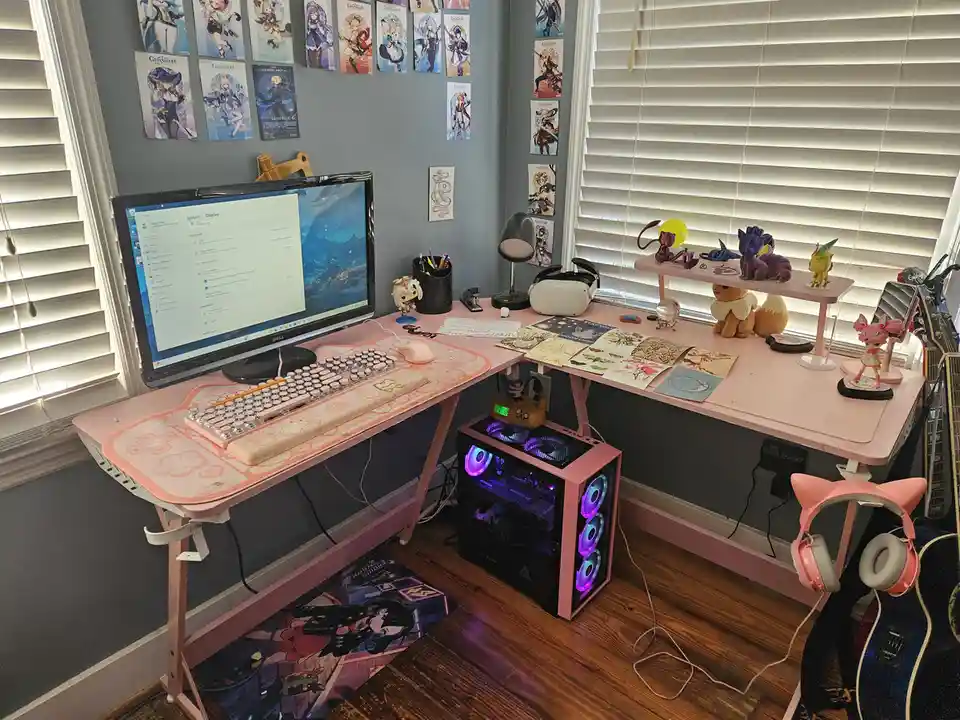 A corner desk setup with a pink computer tower, pink desk, and pink peripherals, surrounded by anime posters and figurines…