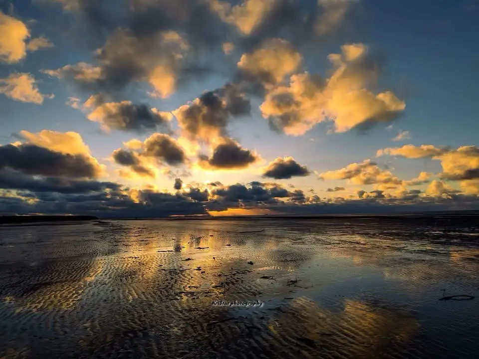 A serene landscape photo of a sunset over a wet, rippled shoreline, with golden clouds reflecting in the water. The overlaid…