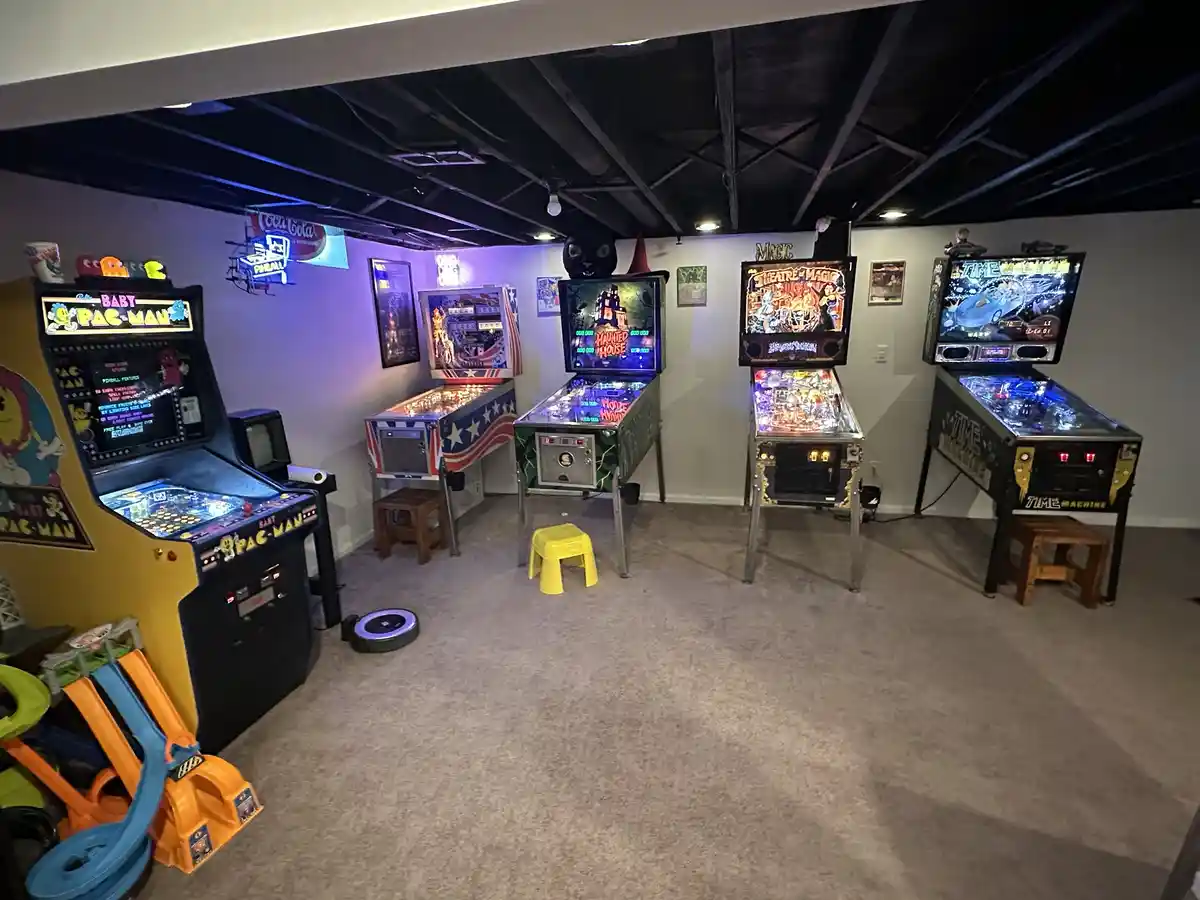 A dimly lit basement room features five vintage arcade and pinball machines, including 'Baby Pac-Man', 'Haunted House'…