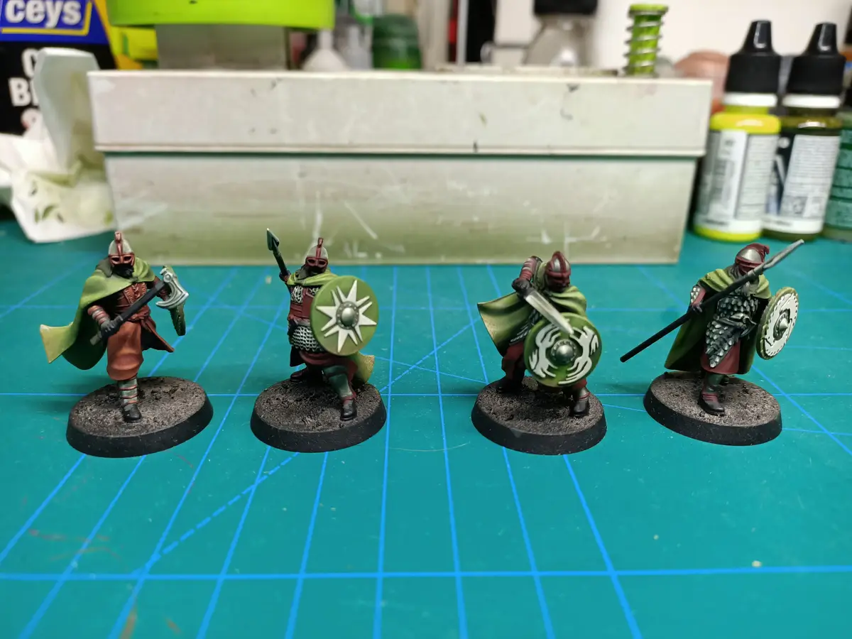 Four painted miniature fantasy warriors in green cloaks and armor stand on circular bases on a green grid mat, each holding…