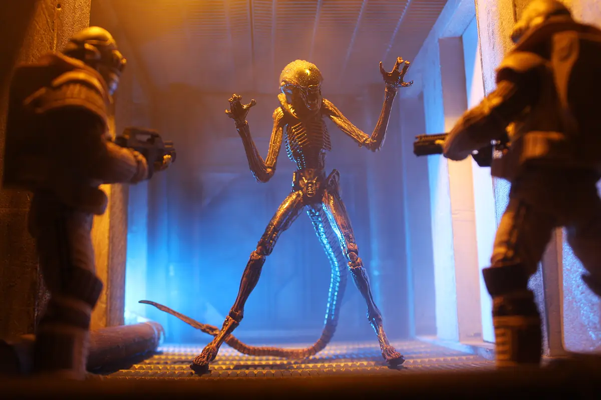A toy alien from the movie 'Alien' stands in a hallway between two toy soldiers holding guns, with dramatic lighting. The…