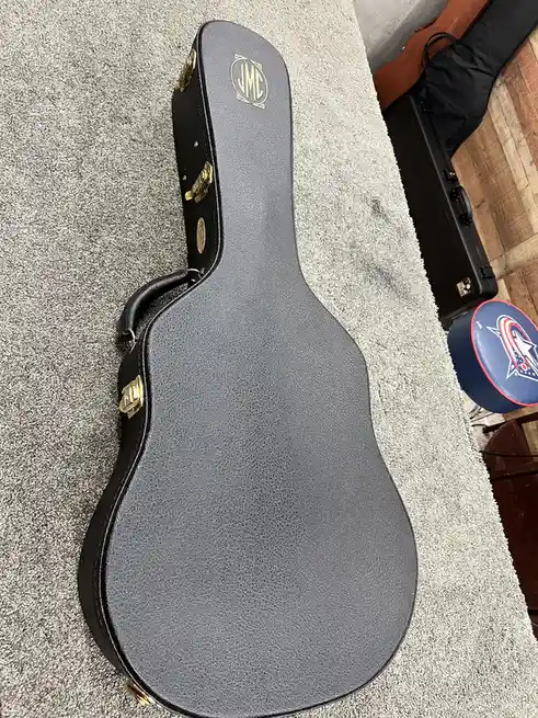 A black, guitar-shaped hard case with gold hardware and a 'JMC' logo lies on a gray carpet. The overlaid text reads 'i…