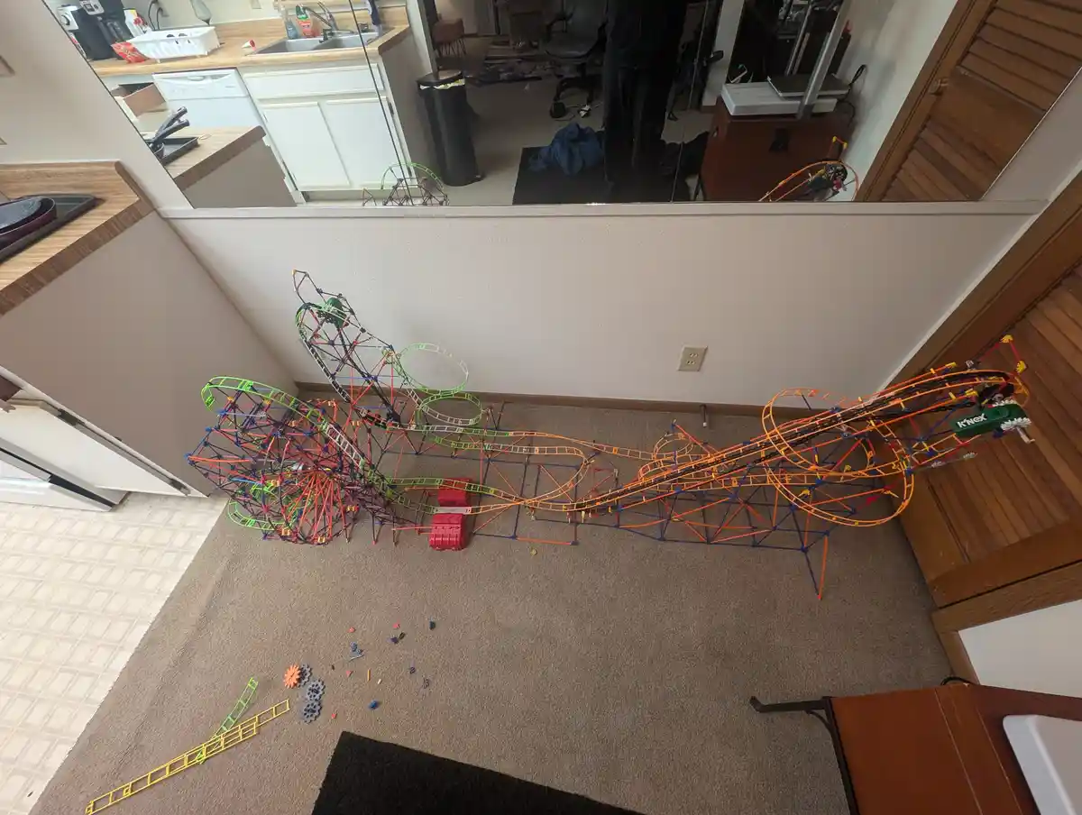 An overhead shot shows a massive, intricate K'nex roller coaster built across a carpeted floor in a living room, with a…