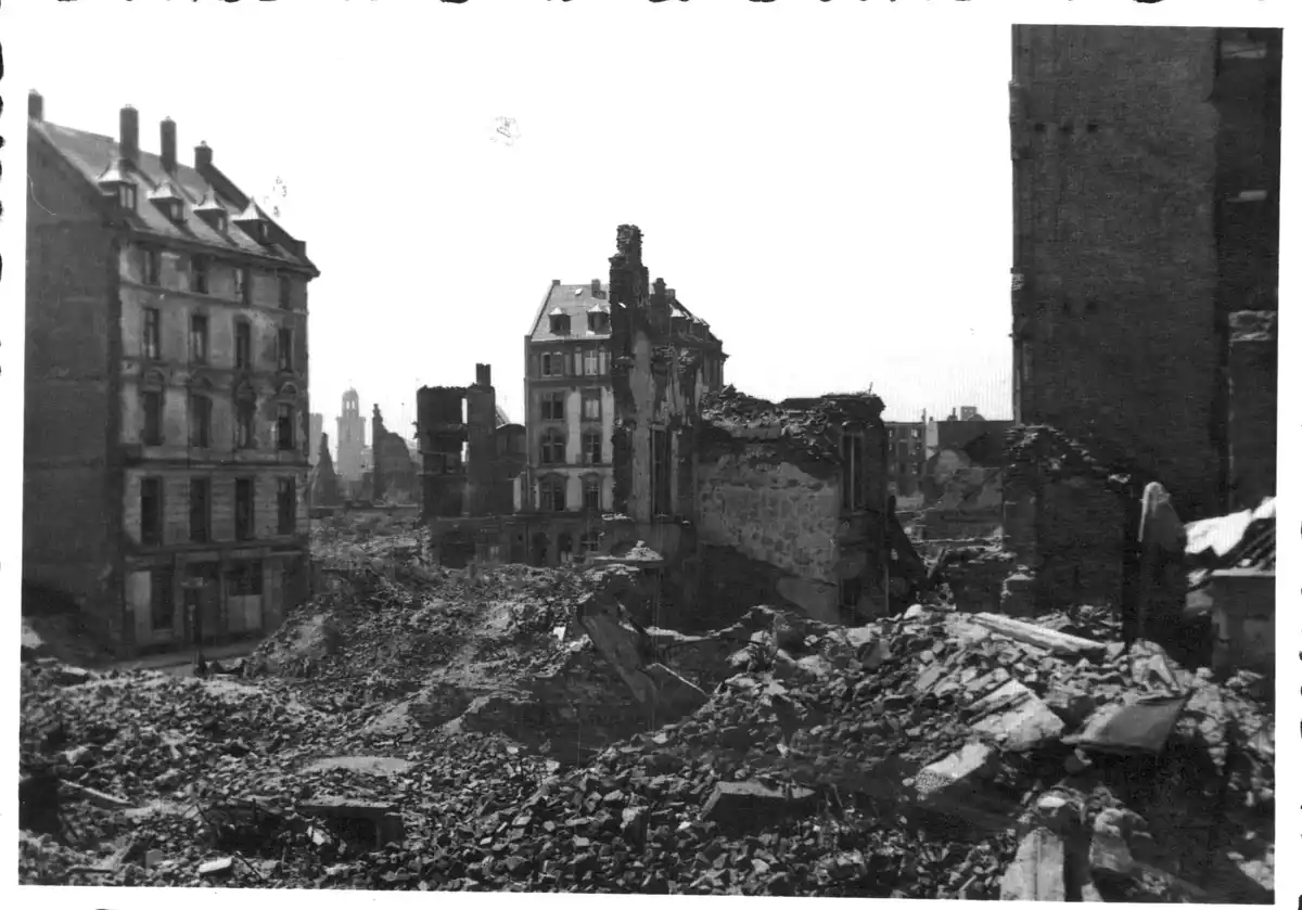A black-and-white historical photograph shows a devastated urban landscape with rubble-strewn streets, partially collapsed…