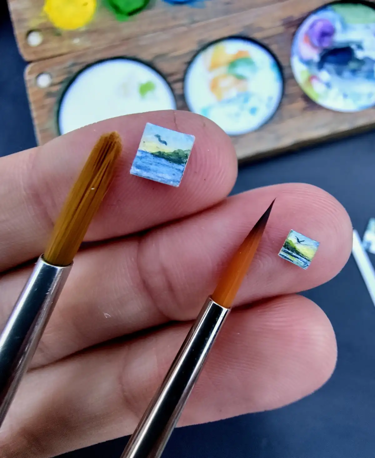 A close-up shot shows two tiny watercolor paintings of landscapes, each resting on a fingertip next to a paintbrush. The…