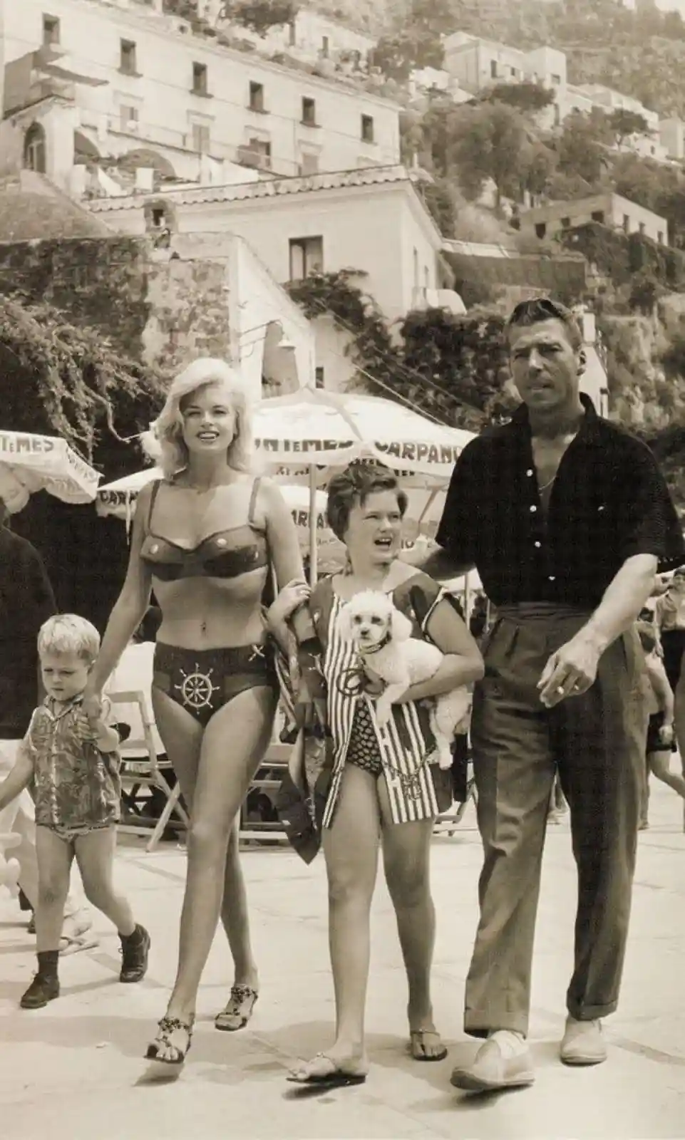 A vintage black-and-white photo shows Marilyn Monroe in a bikini walking with a child, alongside a woman holding a small…