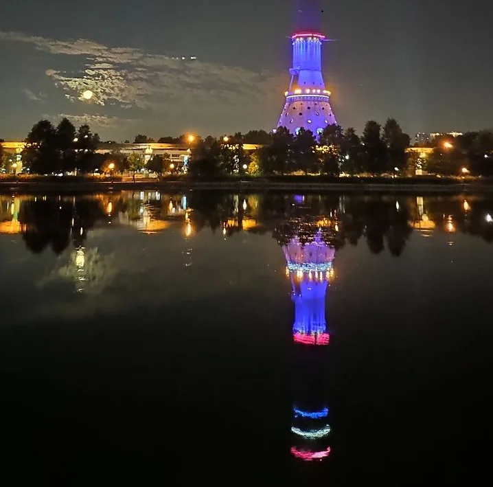 A nighttime photo shows a tall, illuminated tower with blue and red lights, reflected in a calm body of water. The sky above…