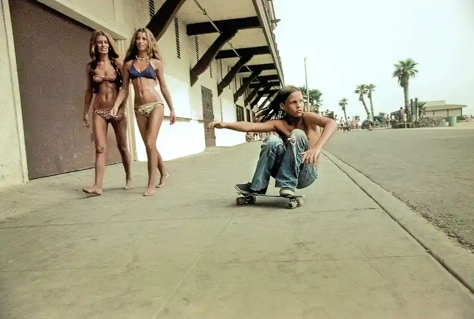 A young person with dreadlocks, wearing ripped jeans and sneakers, crouches on a skateboard with a bored or unimpressed…