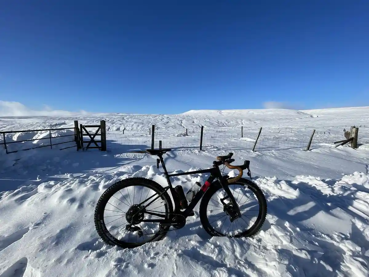 A black bicycle with thick tires and brown handlebars is parked upright in deep snow, facing the viewer. Behind it is a…