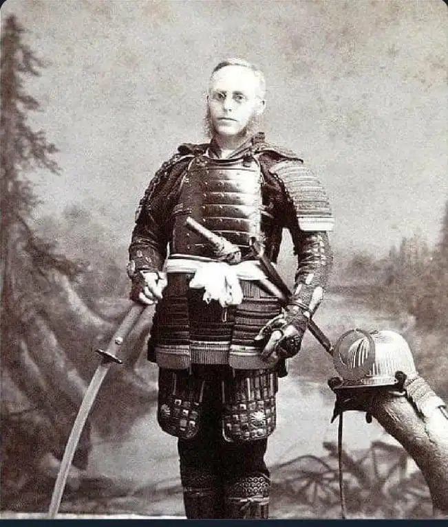 A vintage black-and-white portrait of a man with a thin mustache and glasses, dressed in full traditional Japanese samurai…