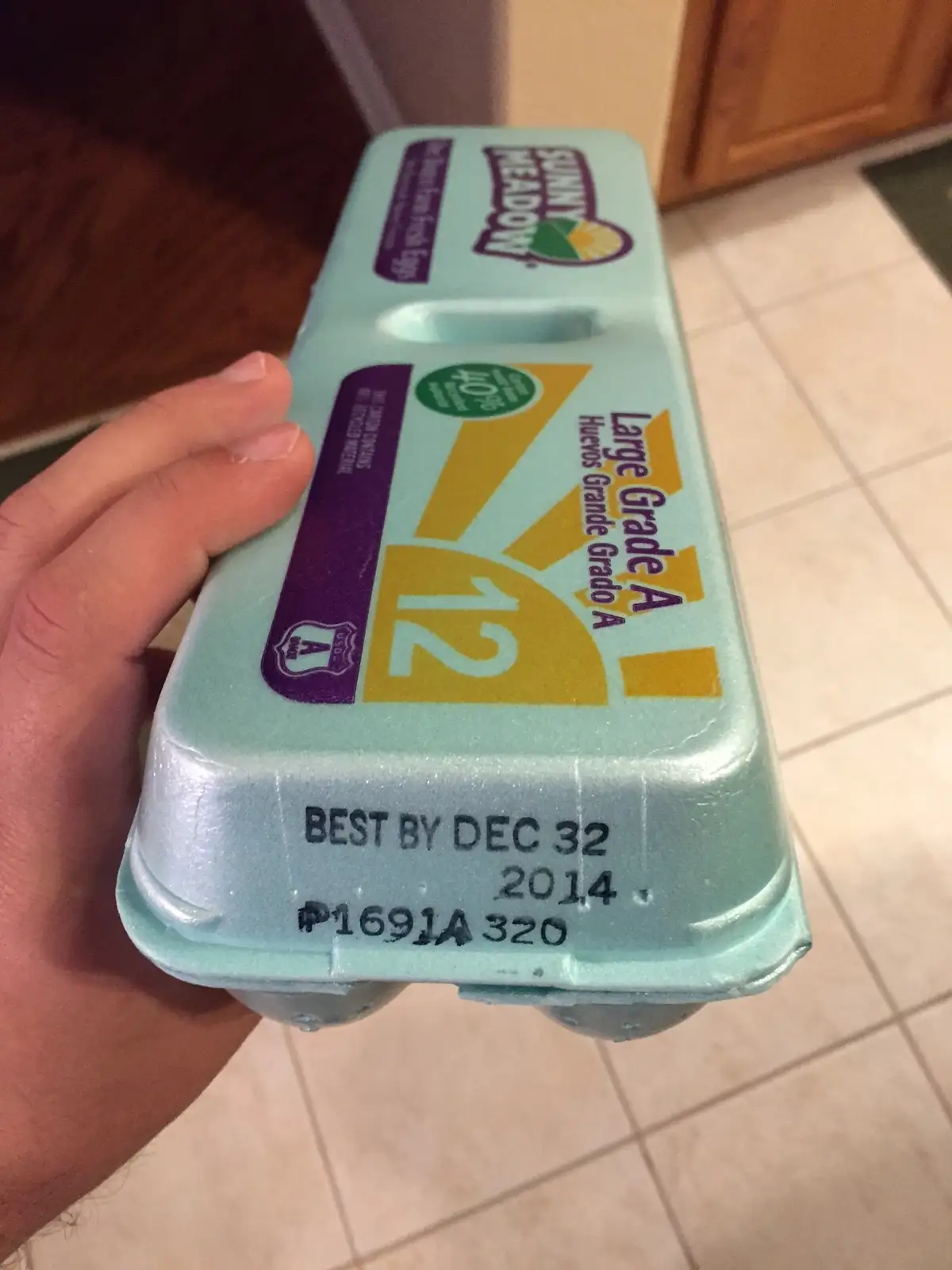 A hand holds a light blue Sunny Meadow egg carton labeled 'Large Grade A' with a 'BEST BY DEC 32 2014' date printed on the…