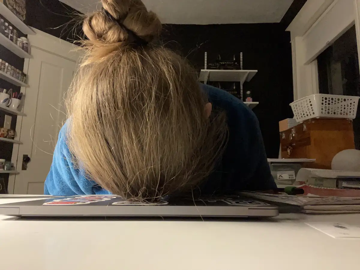 A person with blonde hair in a bun, wearing a blue shirt, is seen from behind with their head resting directly on a laptop…