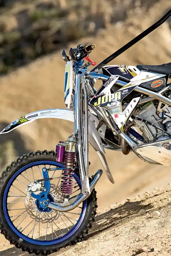 A close-up photo of a dirt bike's front end, showing its chrome frame, purple suspension, blue rim, and various sponsor…