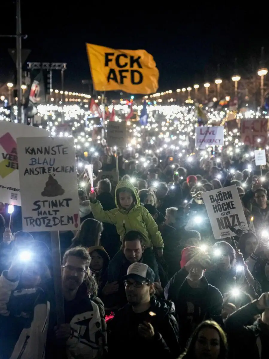 A nighttime protest scene with a large crowd holding up phones with flashlights, creating a sea of light. A child in a…