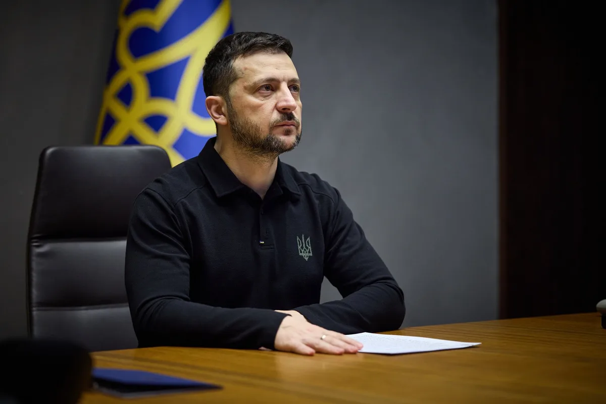 a man with a beard wearing a black polo shirt with a small Ukrainian trident emblem sits at a wooden desk with his hands…