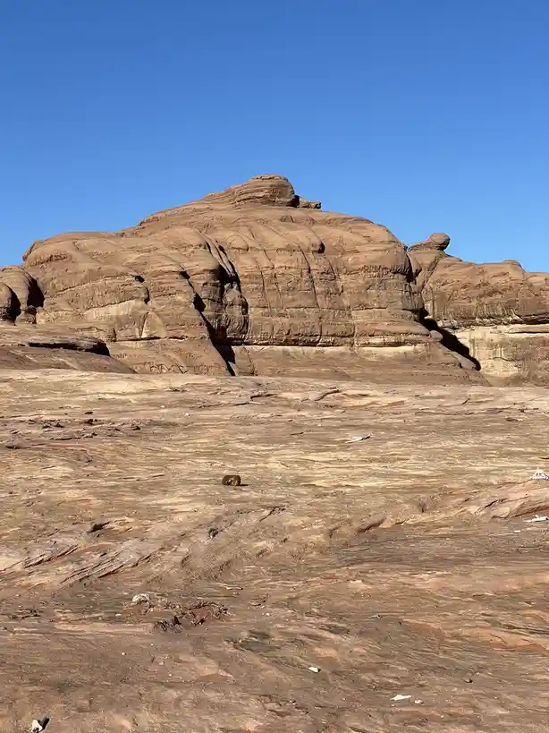 A wide shot of a desert landscape with large, layered sandstone rock formations under a clear blue sky. In the foreground, a…