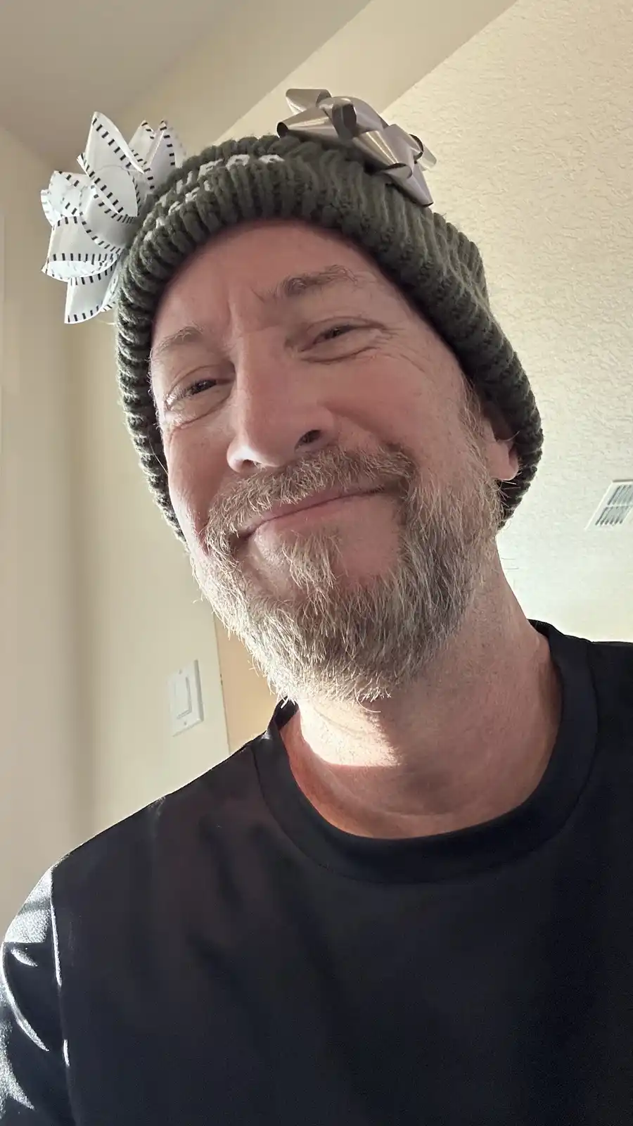 A middle-aged man with a gray beard and mustache smiles while wearing a green knit beanie adorned with two large bows—one…