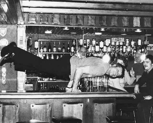 A black-and-white photo shows a man in a bar, balancing horizontally across the bar top with his feet up on the wall and his…