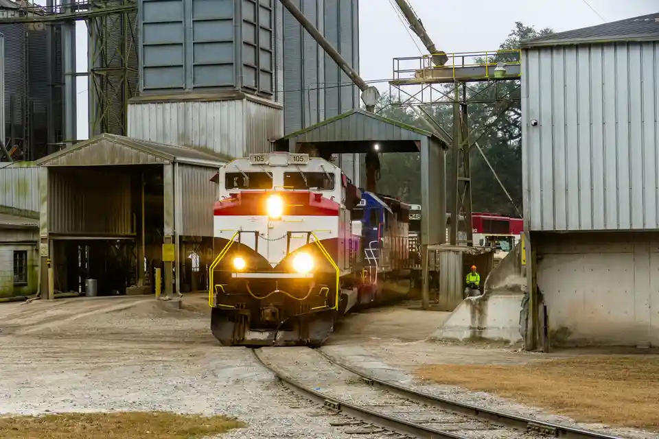 A red and white locomotive with number 105 on its front is approaching on tracks at an industrial rail yard, its headlights…
