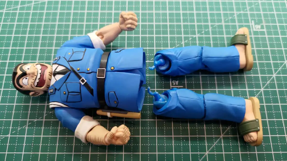 A blue-uniformed action figure, resembling a character from the anime 'Osomatsu-san', lies on its back on a green cutting…
