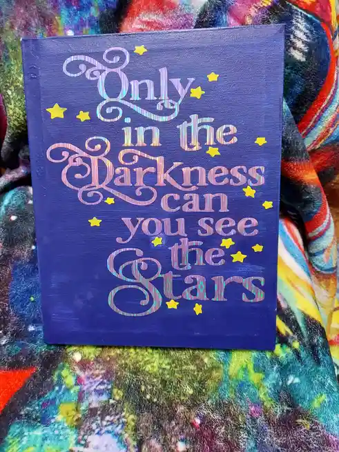 A blue canvas with the phrase 'Only in the Darkness can you see the Stars' written in a shiny, iridescent script font…
