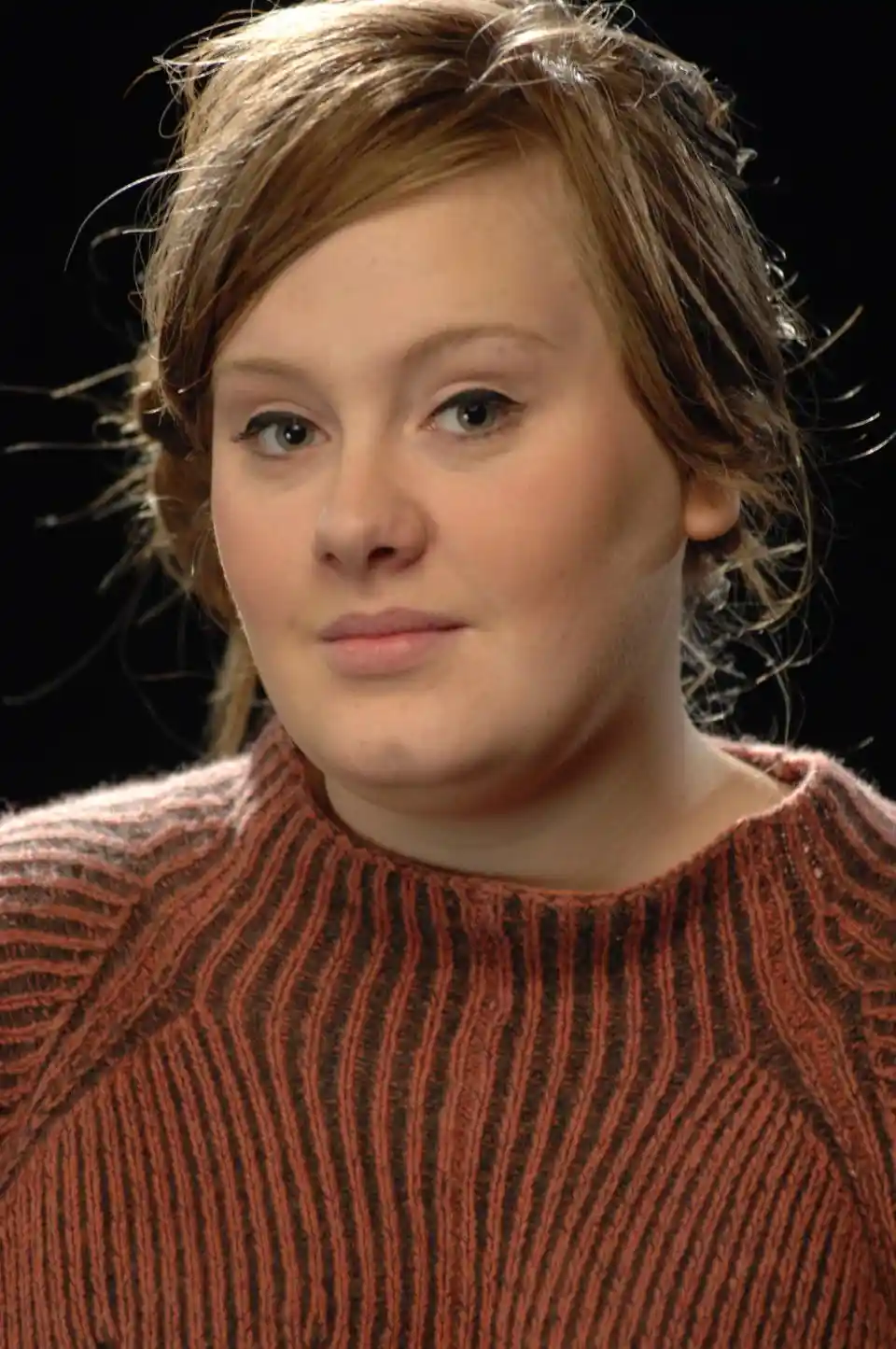 A close-up portrait of a woman with light brown hair and a neutral expression, wearing a rust-colored ribbed sweater against…