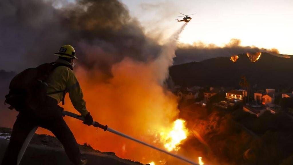 A firefighter in full gear, holding a hose, battles a massive wildfire with flames and thick smoke billowing. In the…