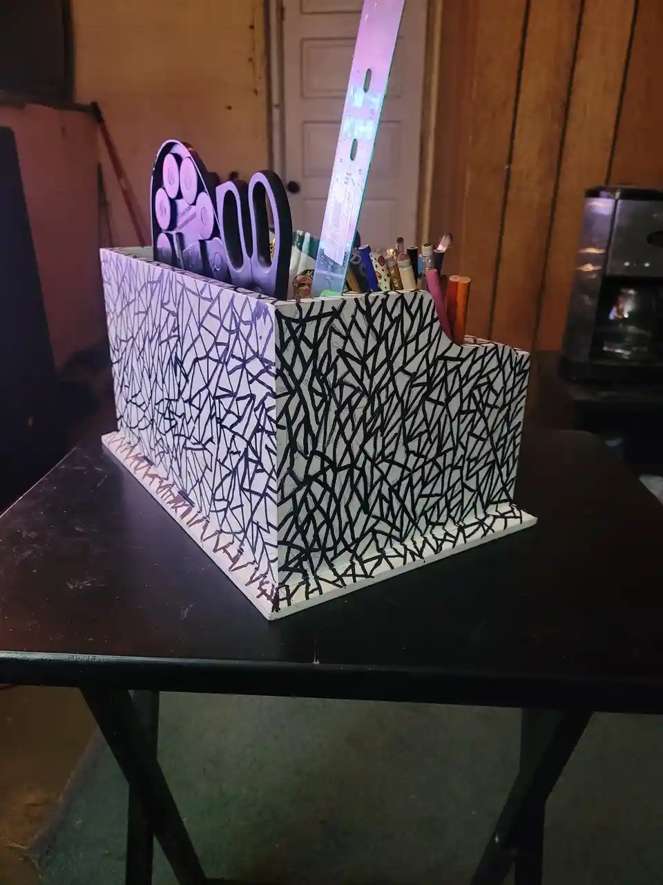 A handmade, angular desk organizer with a black-and-white abstract line pattern sits on a black table, holding scissors, a…