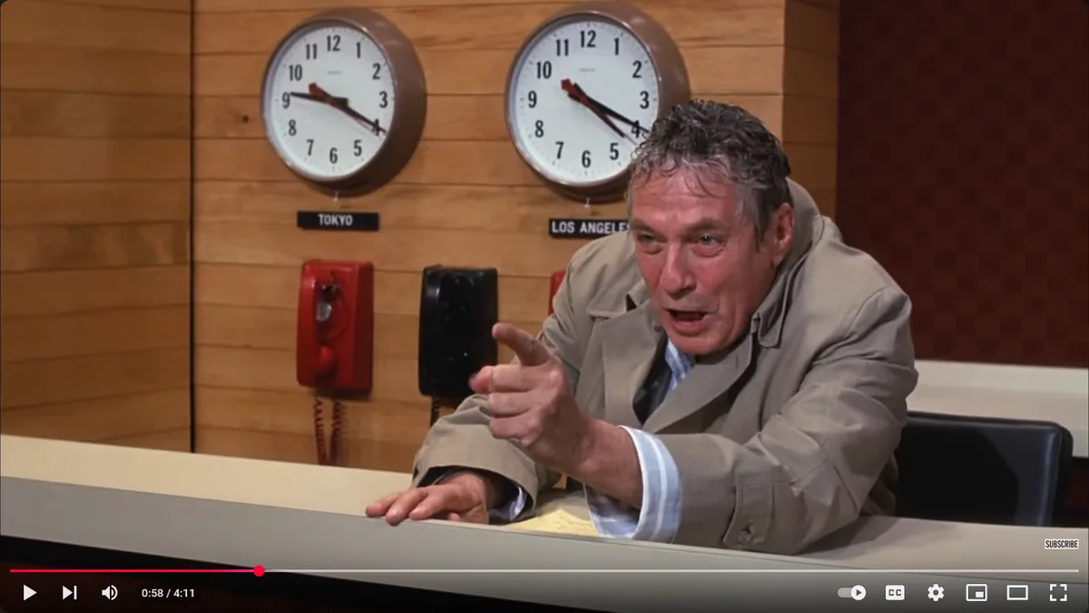 a man in a beige trench coat leans over a desk, pointing angrily at the viewer, with two large clocks labeled 'tokyo' and…